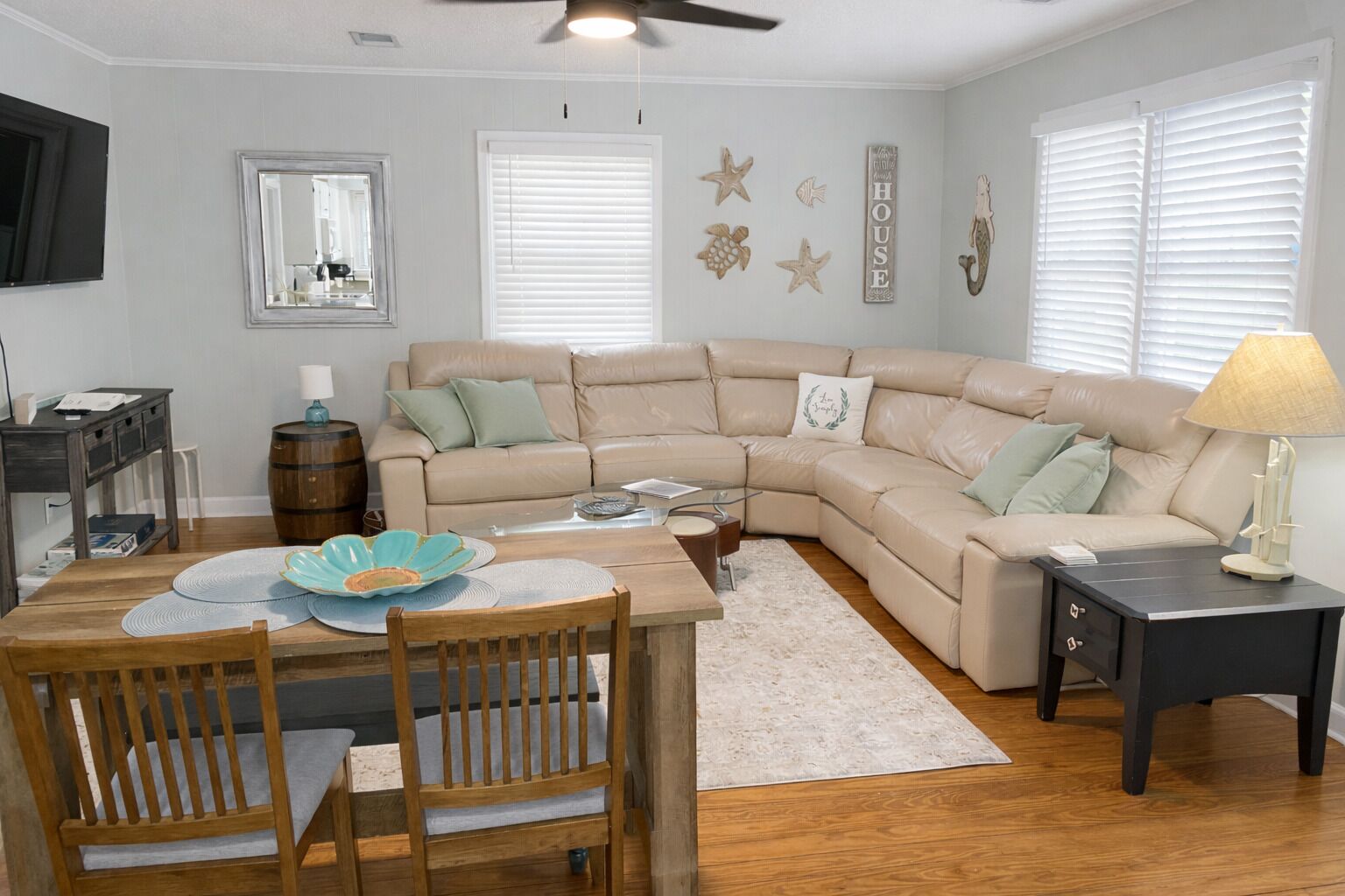 Una sala de estar con un sofá seccional color beige, una mesa de comedor de madera con sillas, una mesa de centro y un televisor montado en la pared.