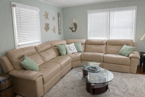 A living room with a beige sectional sofa, a glass coffee table, and decorative wall art. - Relax & Unwind – Ocean Isle Beach Retreat w/ Firepit & Fenced Yard (Ocean Isle Beach)