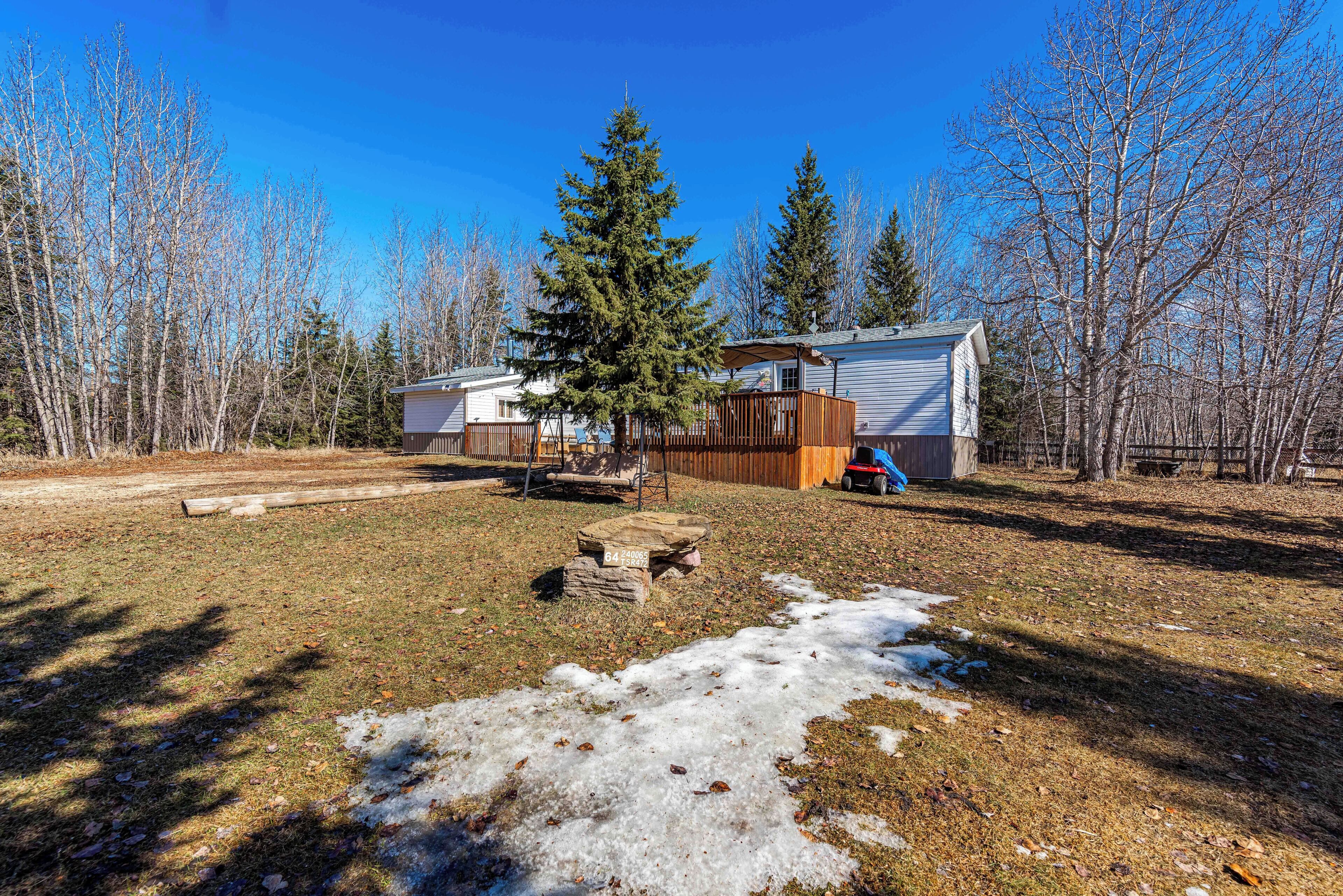 A cabin with a deck, surrounded by trees and a grassy yard with patches of snow.