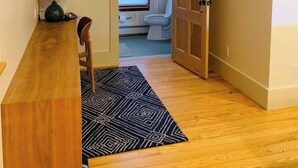En hall som leder till ett badrum med toalett, handfat och fönster. Det finns en trädörr på höger sida, en matta på golvet och en lampa på ett bord till vänster.