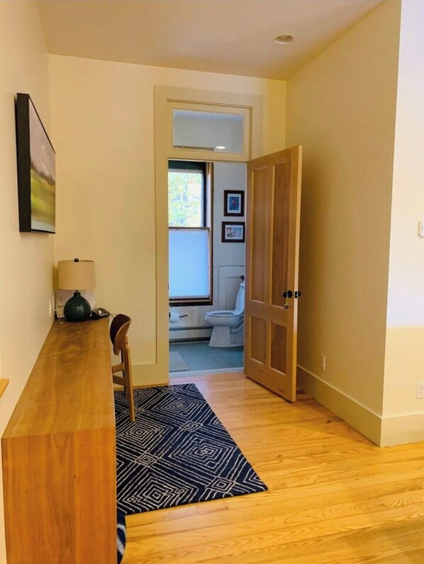 A hallway leading to a bathroom with a toilet, sink, and window. There is a wooden door on the right, a rug on the floor, and a lamp on a table to the left. - Historic, beautifully renovated riverside apartment in North Ferrisburgh (Ferrisburgh)