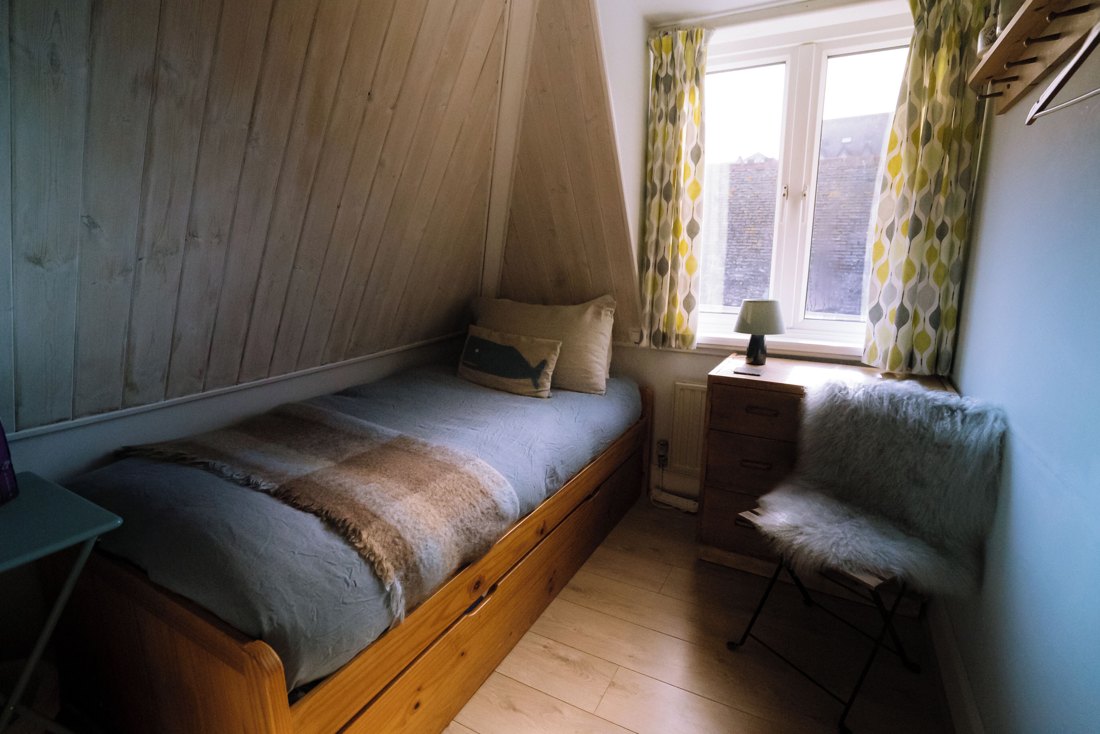 A small, cozy bedroom with a single bed, a chair, a window with curtains, and a radiator.