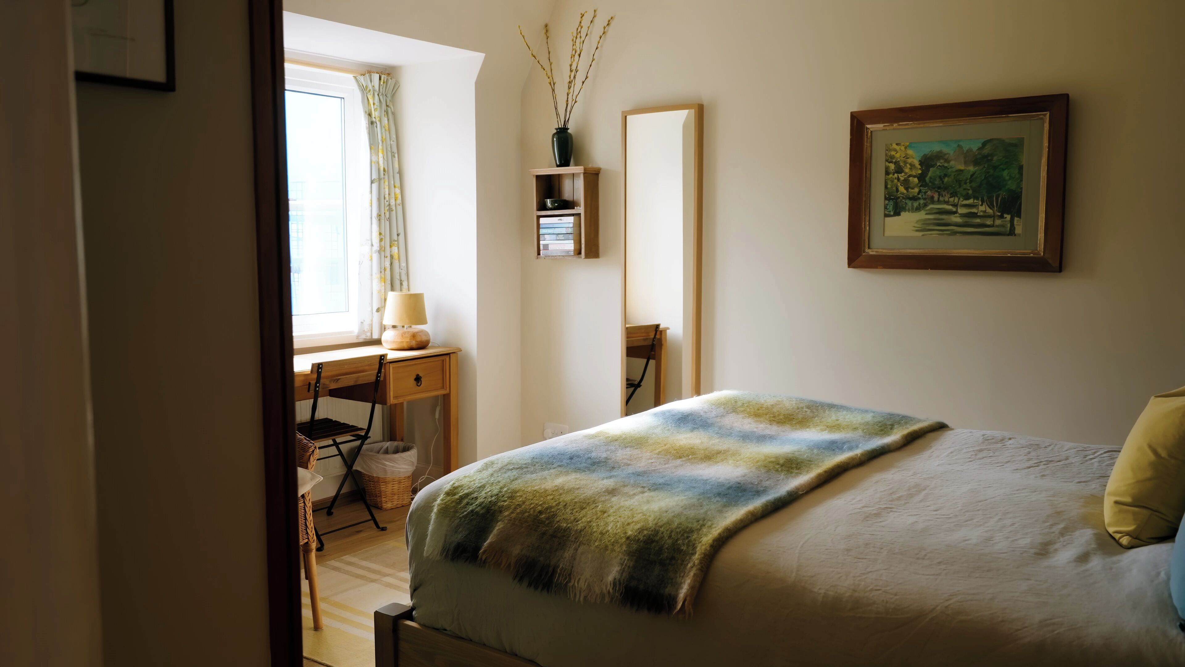 Une chambre à coucher avec un lit, un bureau, une chaise, une fenêtre avec des rideaux, un miroir, un tableau et un vase avec des branches.
