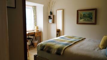 Une chambre à coucher avec un lit, un bureau, une chaise, une fenêtre avec des rideaux, un miroir, un tableau et un vase avec des branches.