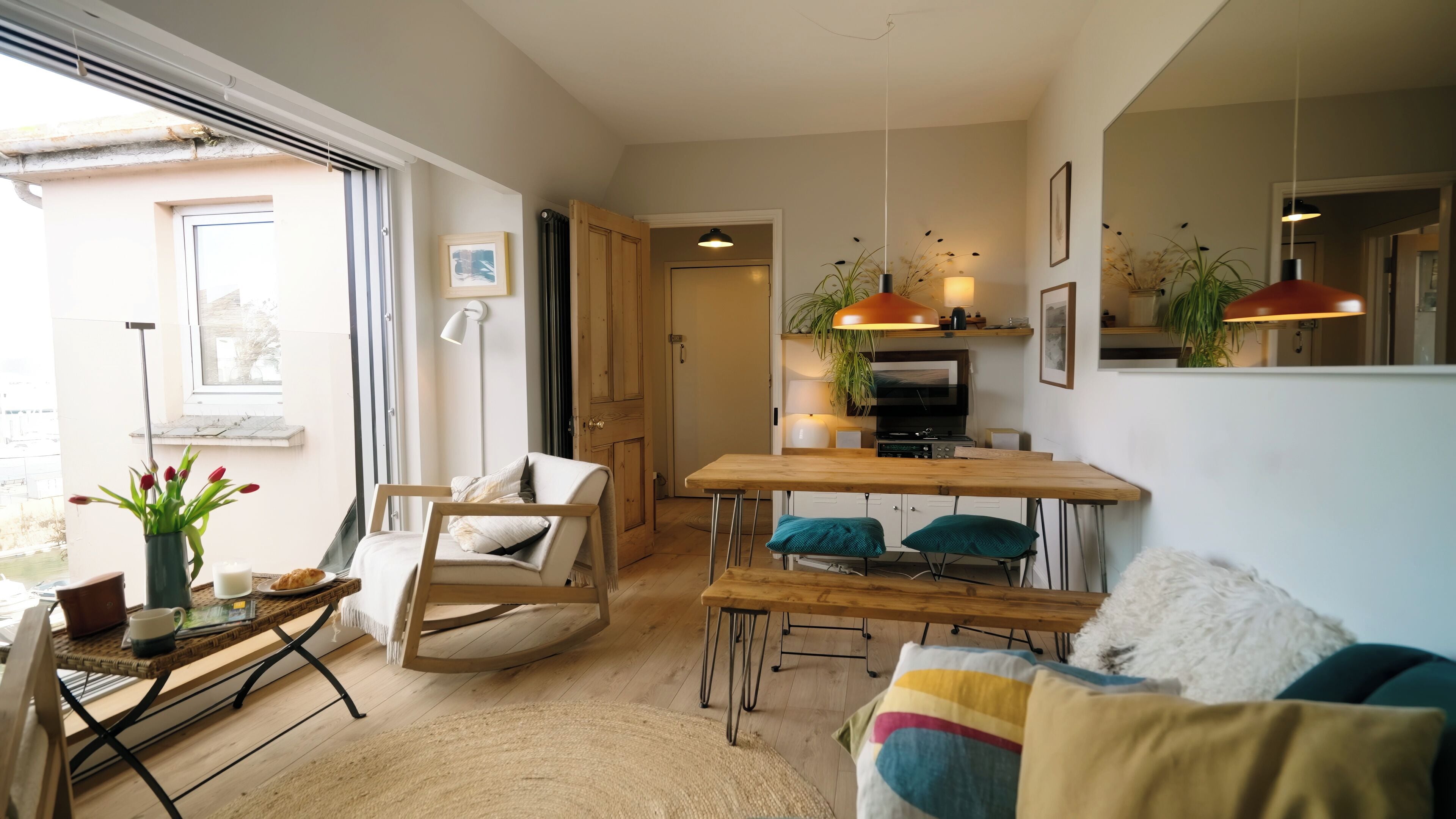 Un salon chaleureux avec une table à manger en bois, un fauteuil confortable et un canapé. Une grande fenêtre laisse entrer la lumière naturelle et offre une vue sur l’extérieur.