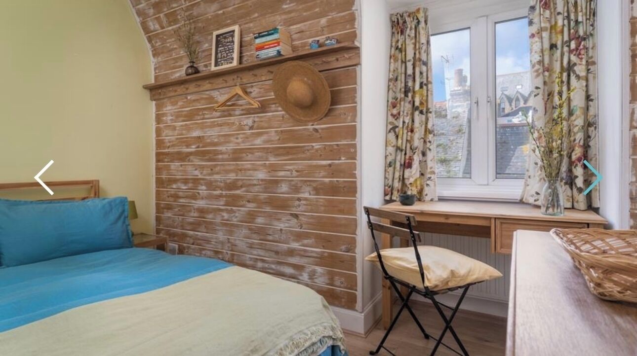 Une chambre avec un mur en bois, un lit avec une literie bleue et beige, un bureau avec une chaise et une fenêtre avec des rideaux.