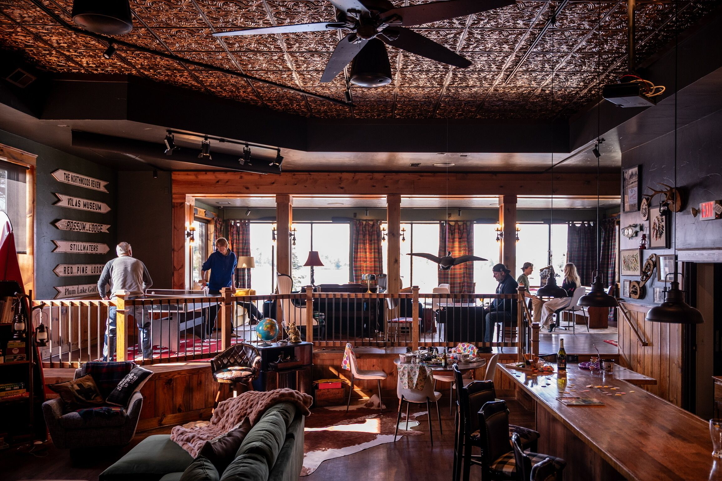 Un área de bar espaciosa con techo alto, vigas de madera y un ventilador grande. Hay varias zonas de estar con mesas y sillas, y una barra con varios objetos sobre ella.