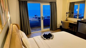 A hotel room with a bed, a desk, a chair, a lamp, and a view of the ocean through a sliding door. - Hotel Midotel Seaview (Pointe-Noire)