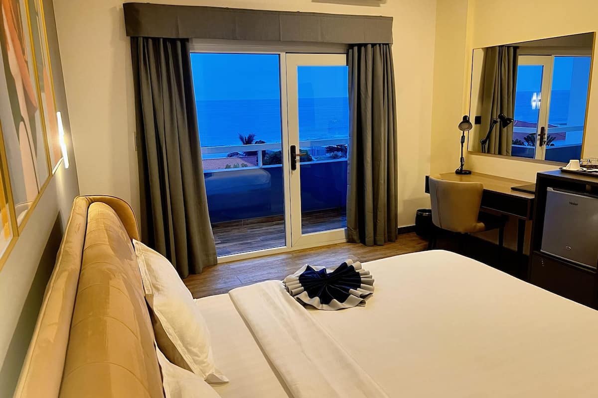 A hotel room with a bed, a desk, a chair, a lamp, and a view of the ocean through a sliding door.