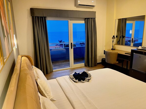 A hotel room with a bed, a desk, a chair, a lamp, and a view of the ocean through a sliding door. - Hotel Midotel Seaview (Pointe-Noire)