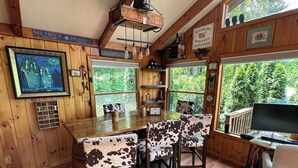 Interior rústico de cabana com mesa de jantar de madeira, cadeiras de couro, área de bar e grandes janelas com vista para o exterior.