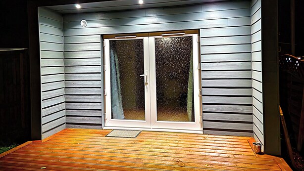 Een overdekt buitengebied met een houten terras, een schuifpui en inbouwverlichting.