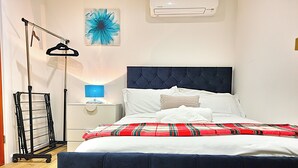 Een net opgemaakt bed met een rood geruit plaid, een donker hoofdbord en een witte nachtkastje met een blauwe lamp.