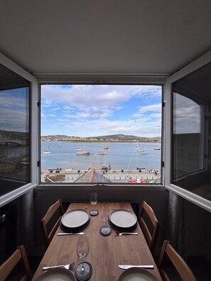 Comedor con una mesa dispuesta para cuatro, con vistas a un puerto con barcos y una ciudad al fondo.