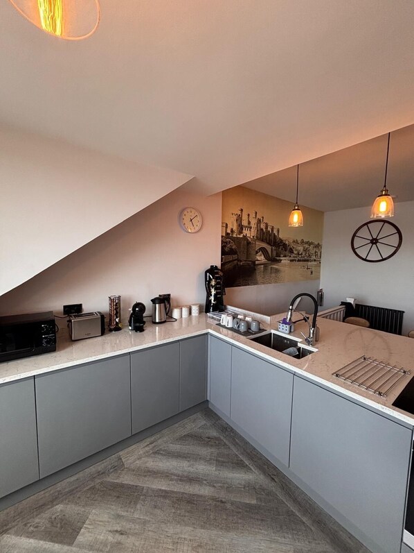 Una cocina moderna con gabinetes grises, piso con patrón de espiga, un reloj, una pintura de paisaje urbano, lámparas colgantes y un hervidor.