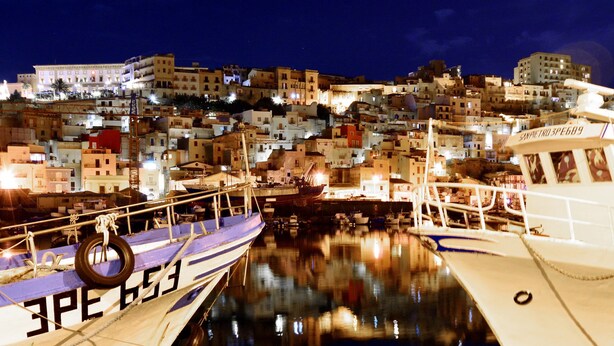 A harbor with boats docked, a building with multiple stories, and a cityscape in the background.