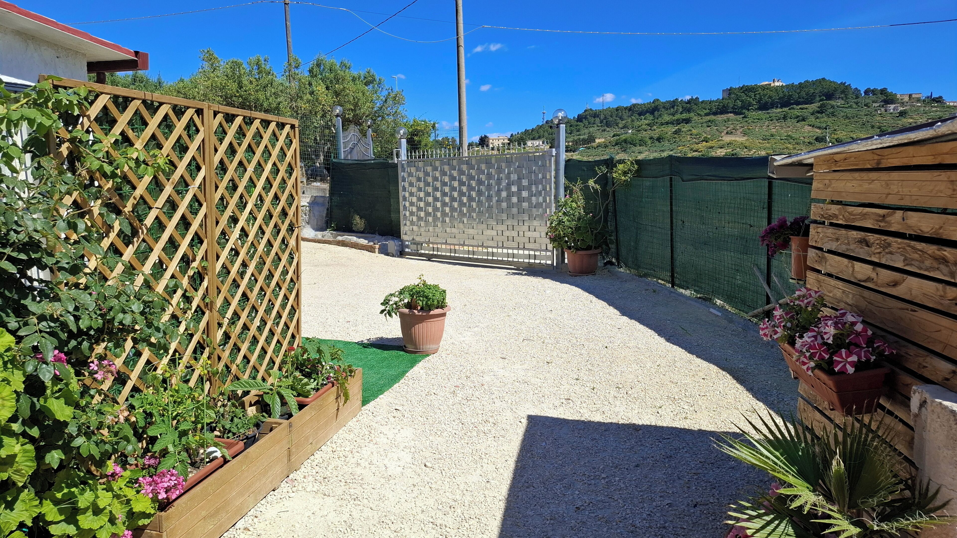 Un chemin de gravier mène à une entrée fermée par un portail, qui est elle-même entourée de clôtures en bois et de plantes en pot.