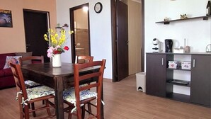 A dining area with a wooden table and chairs, a vase with flowers, and a kitchen area with appliances and shelves.