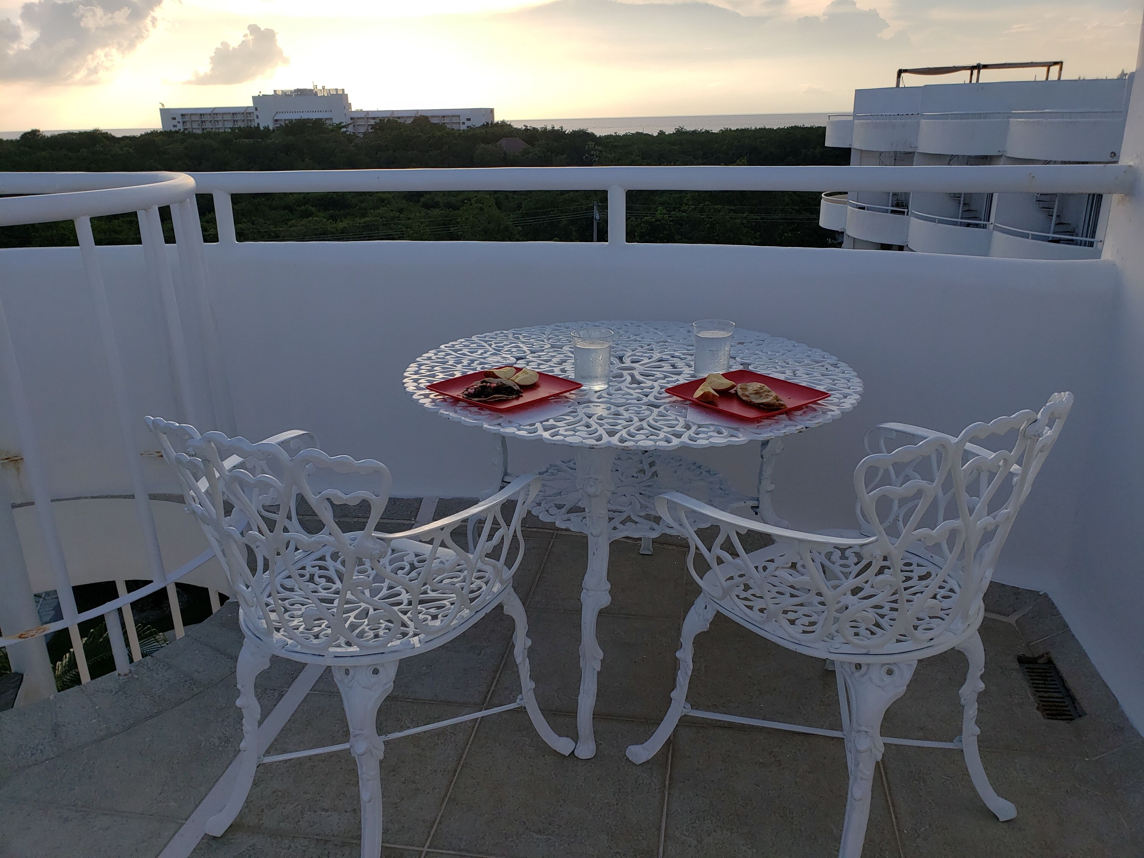 A white outdoor dining area with a round table and chairs, overlooking a cityscape and greenery.