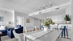 A modern living room with a white sofa, blue armchairs, a dining table with white chairs, and a kitchen area with white cabinets and a plant. - Boardwalk to Beach ready! Boat Slip & Parking. (Ocean City)