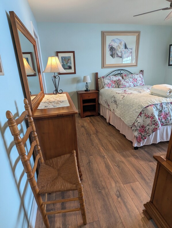 Un dormitorio con una silla de madera, una cama con ropa de cama floral, una mesita de noche con una lámpara y cuadros enmarcados en la pared.