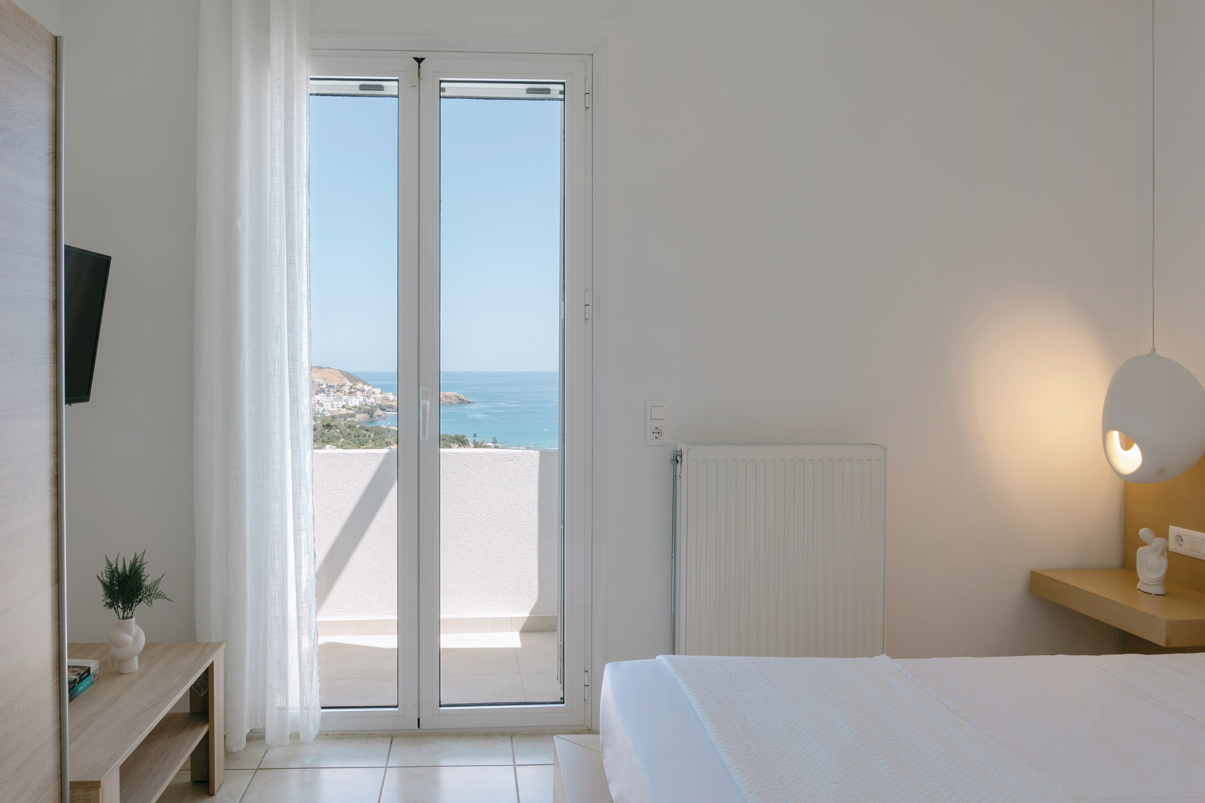 Un dormitorio con cama, mesita de noche, lámpara, televisor en la pared y vista al mar a través de una puerta corrediza.