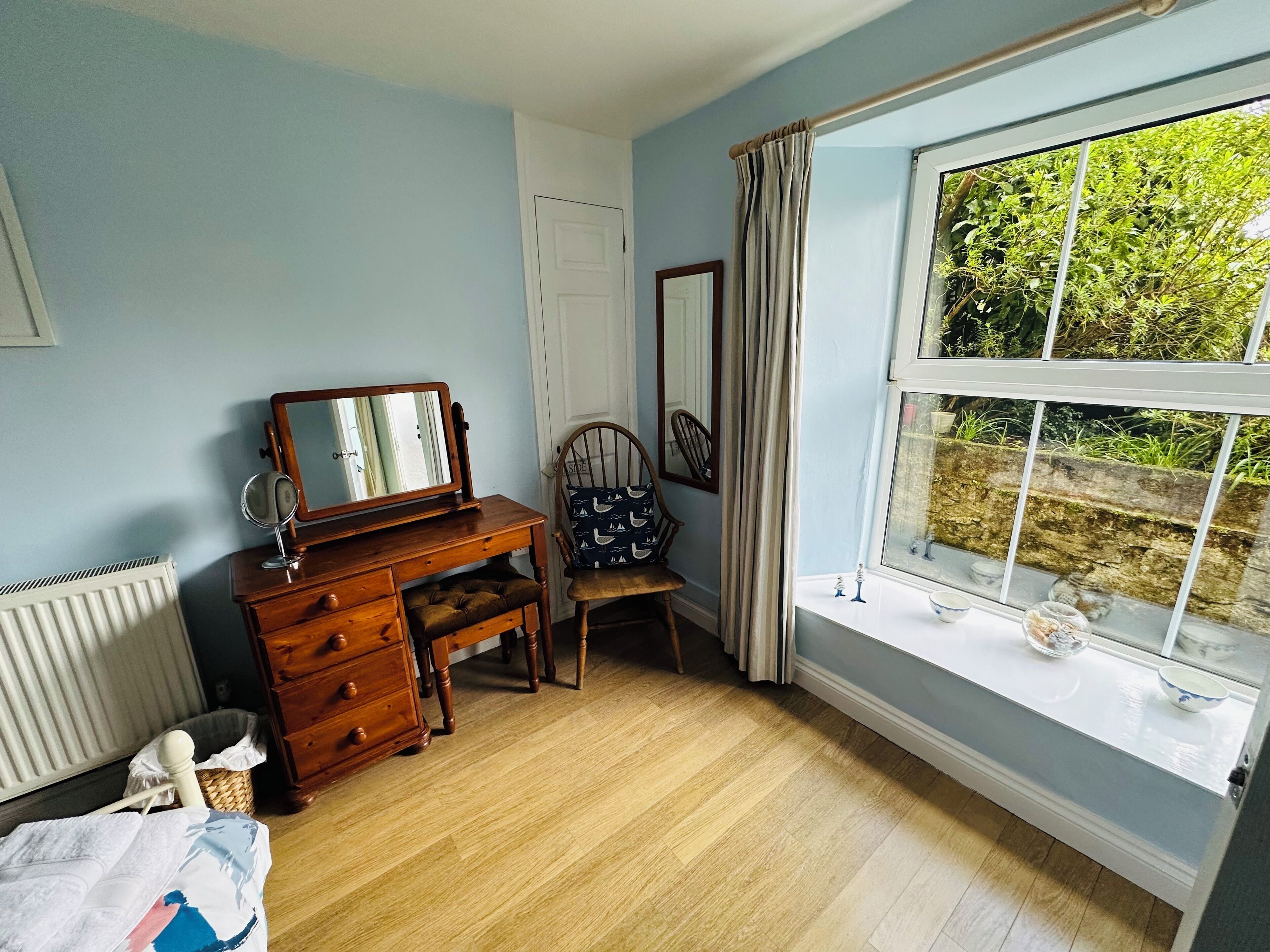 Een kamer met een houten dressoir, een stoel, een raam met uitzicht op groen en een radiator.