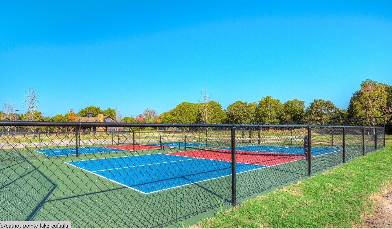 A tennis court with a chain-link fence, surrounded by grass and trees.