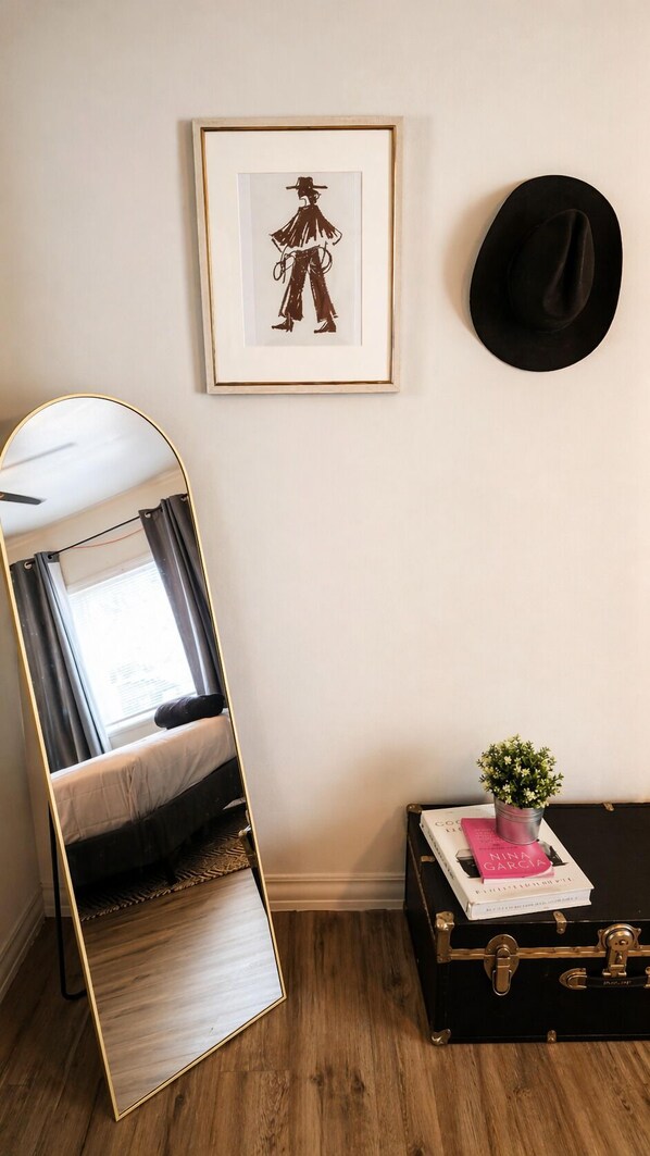 A bedroom with a bed, a large mirror, a black hat, a framed picture, and a suitcase with a plant.
