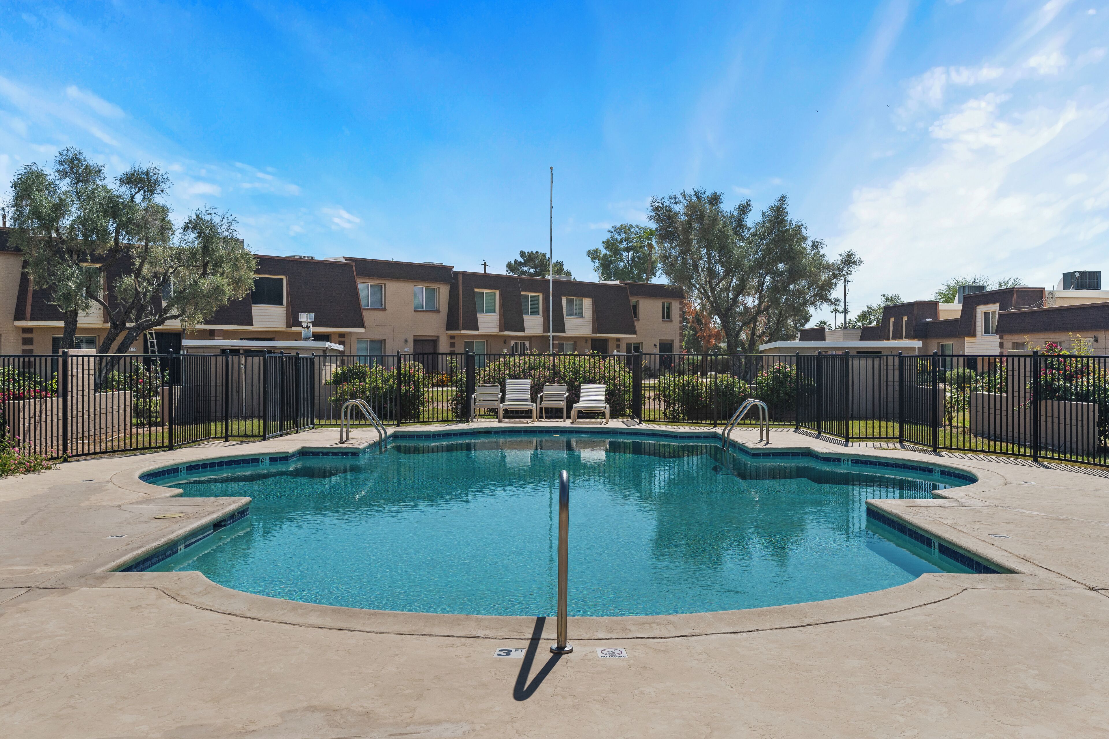 A swimming pool with a metal railing, surrounded by a fence. There are lounge chairs and a basketball hoop in the background.