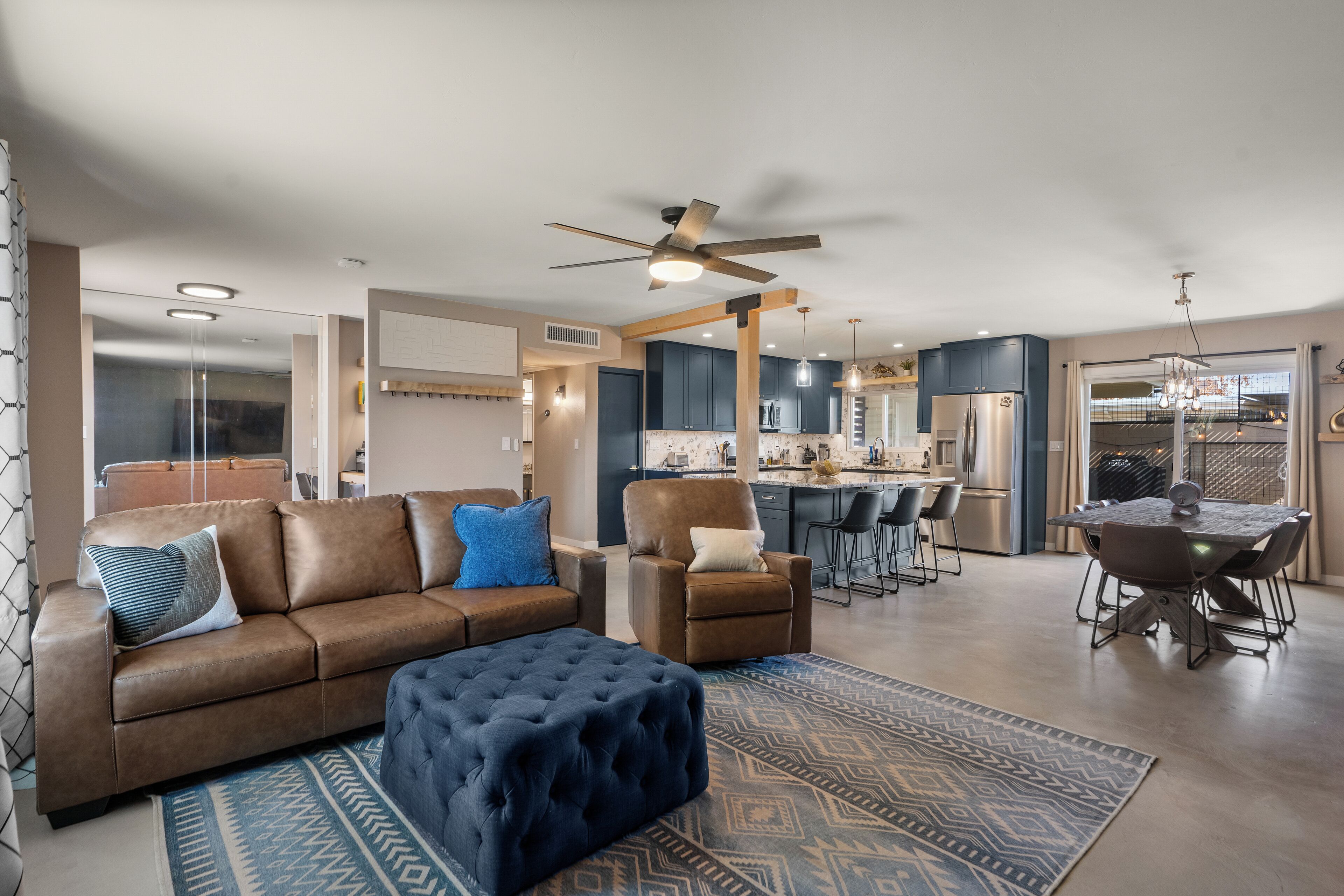 A spacious living room with a brown leather sofa, a blue ottoman, and a dining area with a table and chairs.