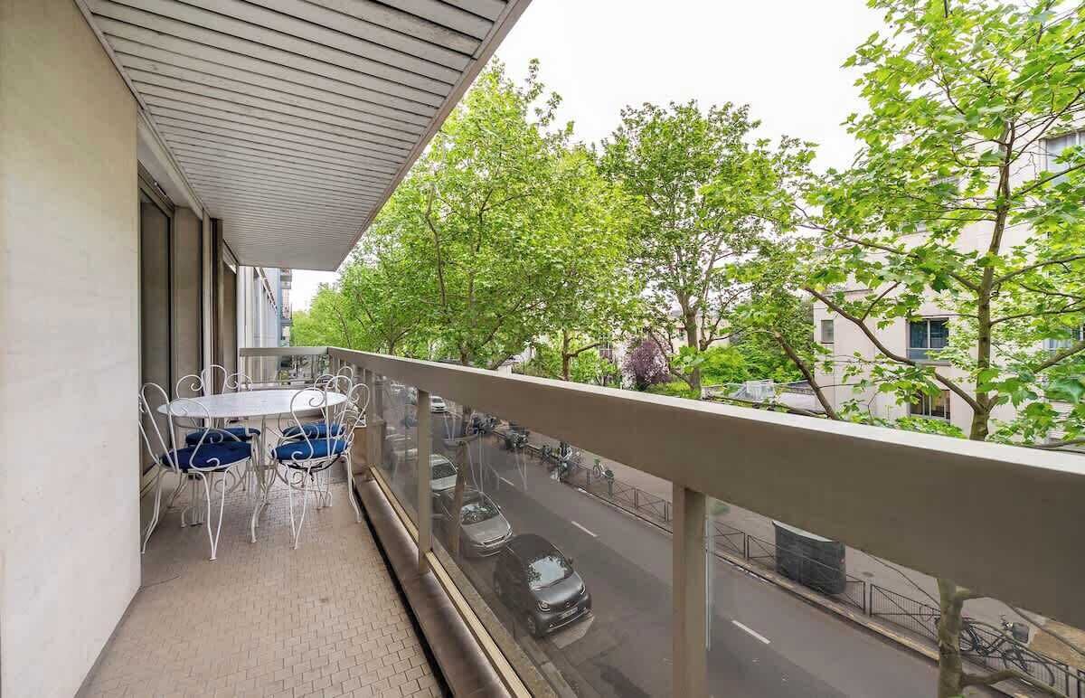 Un balcon avec une table et des chaises, donnant sur une rue bordée d’arbres et où circulent des voitures.