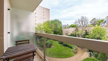 Een balkon met een houten bankje, een glazen hek en uitzicht op een binnenplaats met groen en gebouwen.