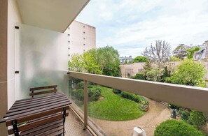 Een balkon met een houten bankje, een glazen hek en uitzicht op een binnenplaats met groen en gebouwen.