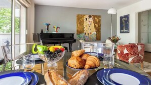 A dining area with a piano, a painting, and a table set with croissants, fruit, and glasses.