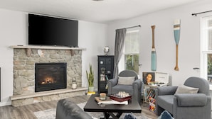 A living room with a stone fireplace, a flat-screen TV, a grey armchair, a coffee table with chess pieces, and a bookshelf.