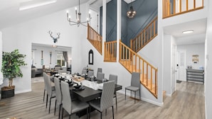 A spacious dining area with a high ceiling, a staircase with wooden railings, and a modern kitchen in the background.
