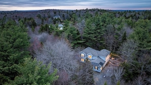 A two-story house surrounded by dense forest, with a visible driveway and a bench outside.