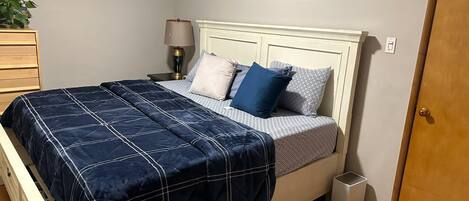 Una cama bien hecha con un edredón azul, una lámpara de noche y una obra de arte en la pared.