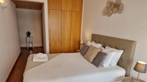 Ein ordentlich bezogenes Bett mit weißer Decke und mehreren Kissen, ein Nachttisch mit Lampe sowie ein Holzschrank im Hintergrund.