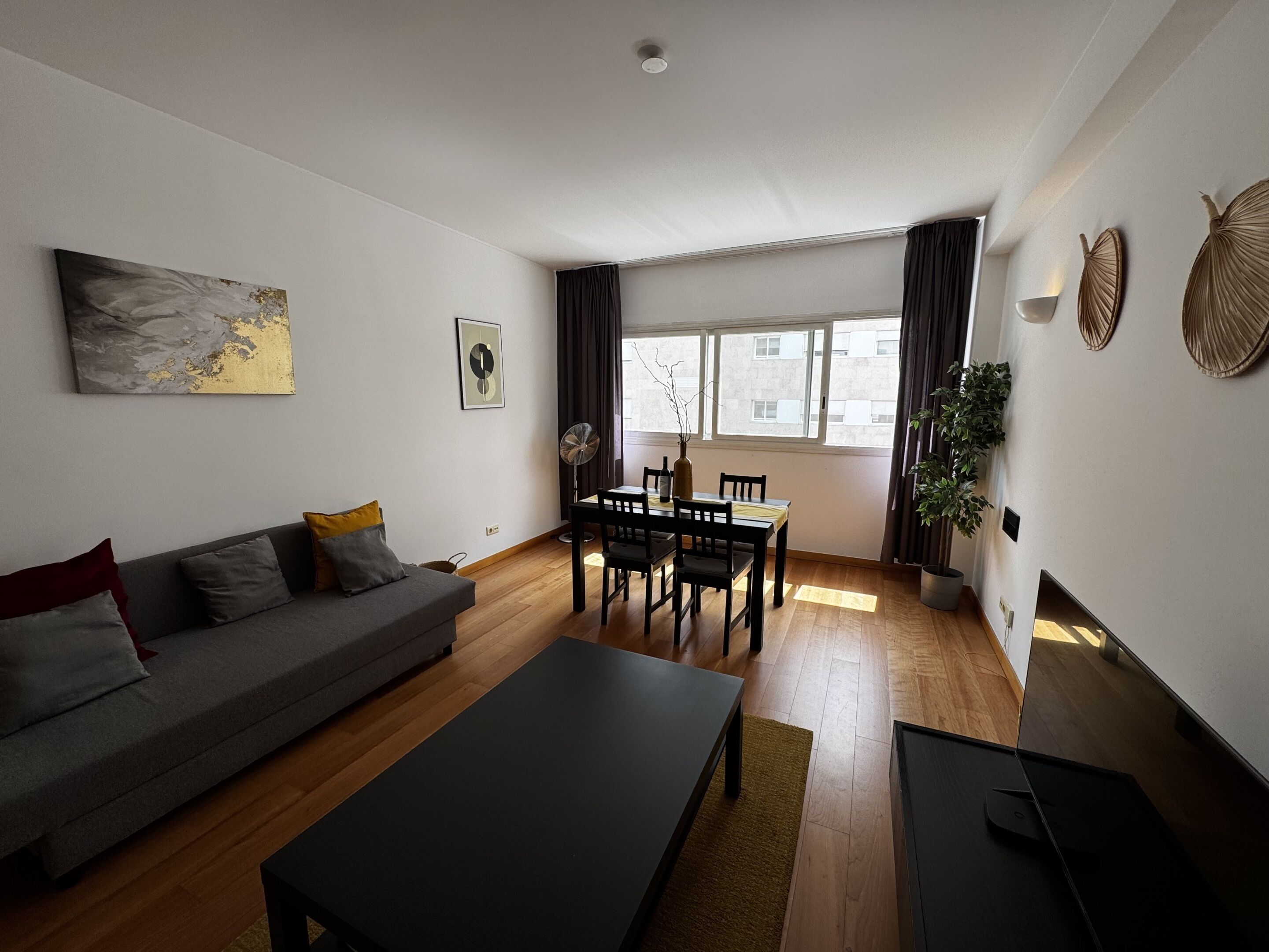 A modern living room with a sofa, a dining table, and chairs. There is a large window with curtains, a fan, and a potted plant.