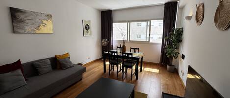 A modern living room with a sofa, a dining table, and chairs. There is a large window with curtains, a fan, and a potted plant.