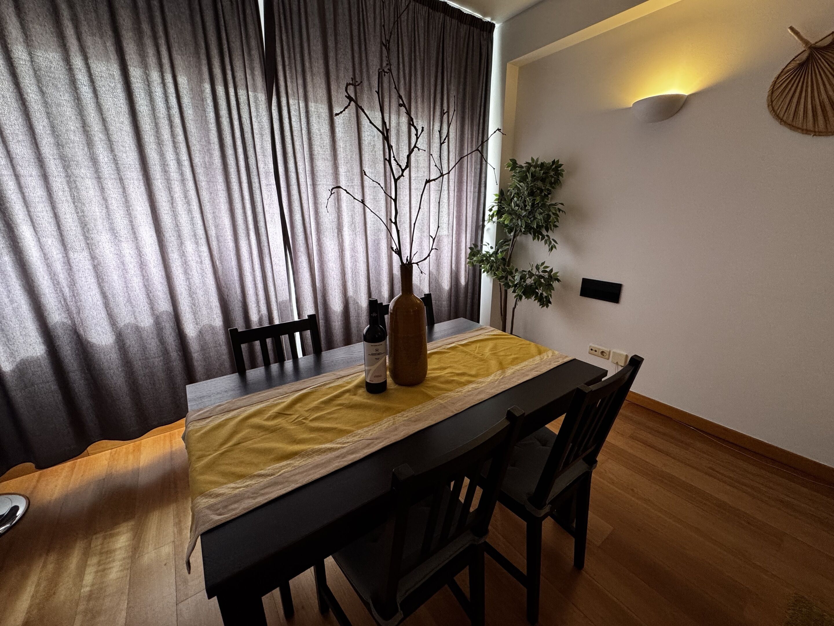 Et spiseområde med et bord dækket til to personer, en vase med grene og en potteplante.
