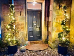 A dark wooden door with the number 9, flanked by two potted plants with string lights, set against a stone wall. - Ladies Lodge - Inverness (Inverness)