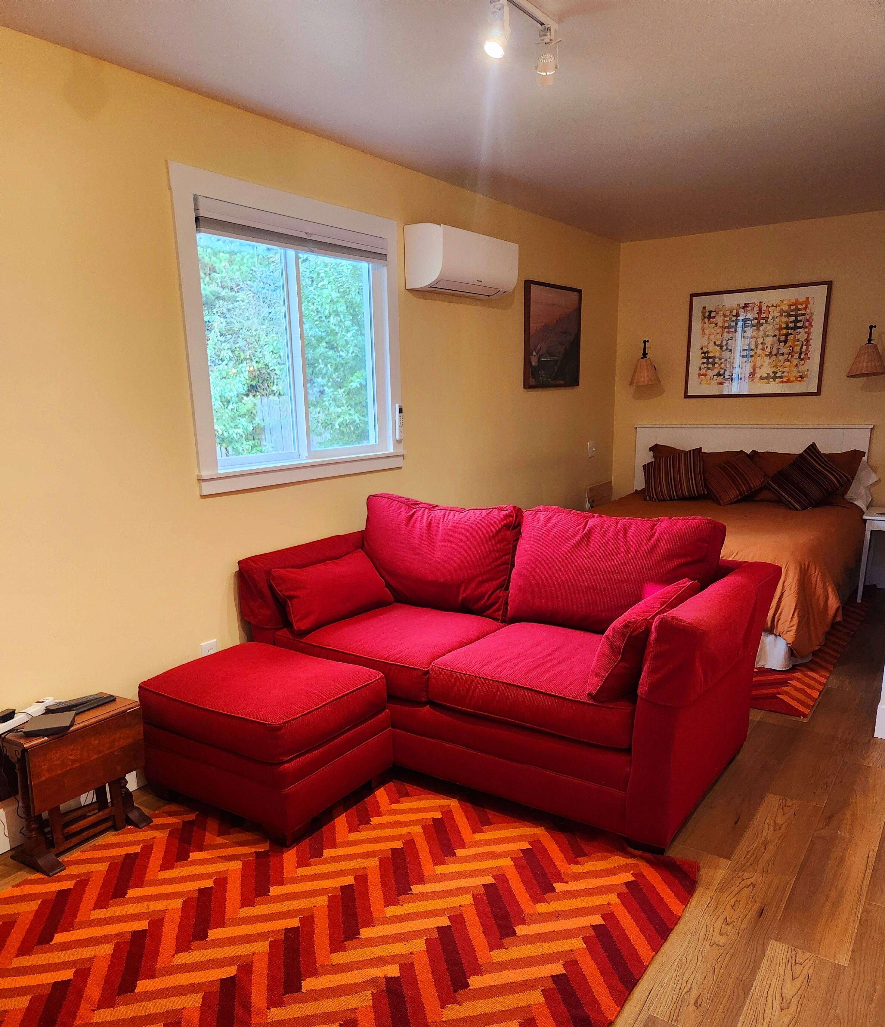 Quarto com sofá seccional vermelho, cama com roupa de cama marrom, janela com persianas, unidade de ar condicionado e tapete com padrão herringbone.
