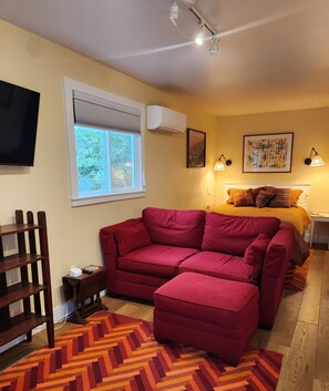 Une pièce chaleureuse avec un canapé rouge, une chaise en bois, un lit recouvert d’une couverture jaune, une fenêtre avec des stores et un téléviseur fixé au mur.