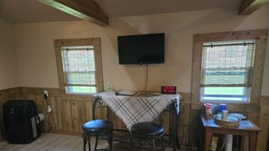 Habitació amb parets de fusta, un televisor fixat a la paret, dos tamborets, una taula amb mantell a quadres i un rellotge que marca les dotze.