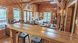 A wooden cabin interior with a kitchen area, a dining table, and a living room with a sofa and chairs.