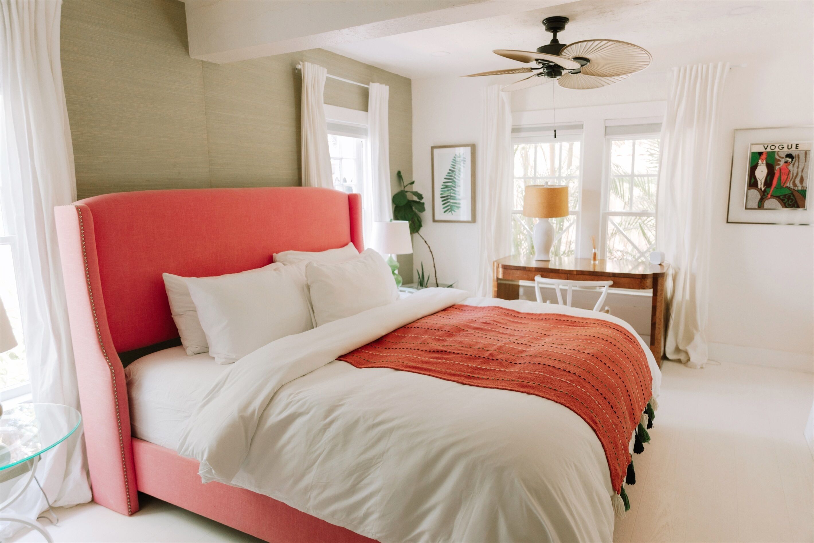 A bedroom with a pink bed, white bedding, a desk with a lamp, and a ceiling fan.