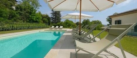 A swimming pool with lounge chairs and umbrellas, surrounded by greenery and a building.